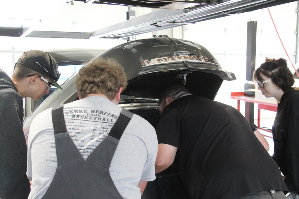 Auto service teacher shows students underneath the hood of a vehicle.