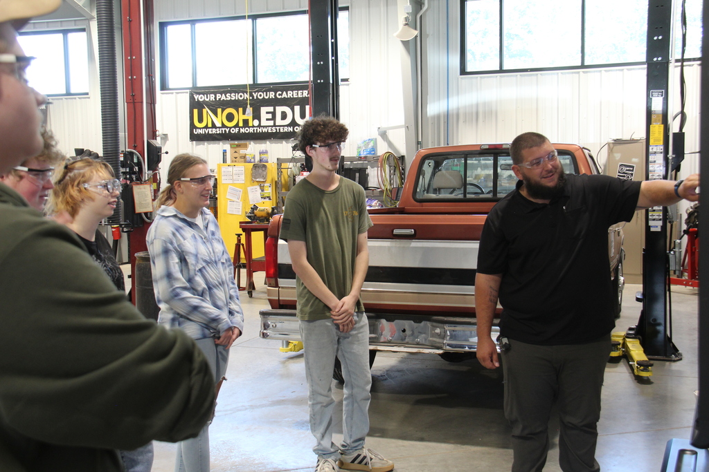 Auto Service teachers points at the monitor as students stand watching to figure out their day. 