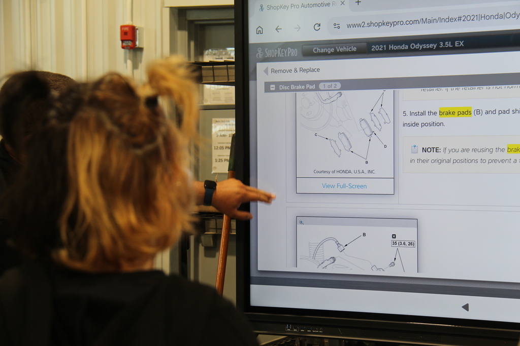 Students stands listening as the teacher points to the monitor showing them how to remove and replace a brake pad. 