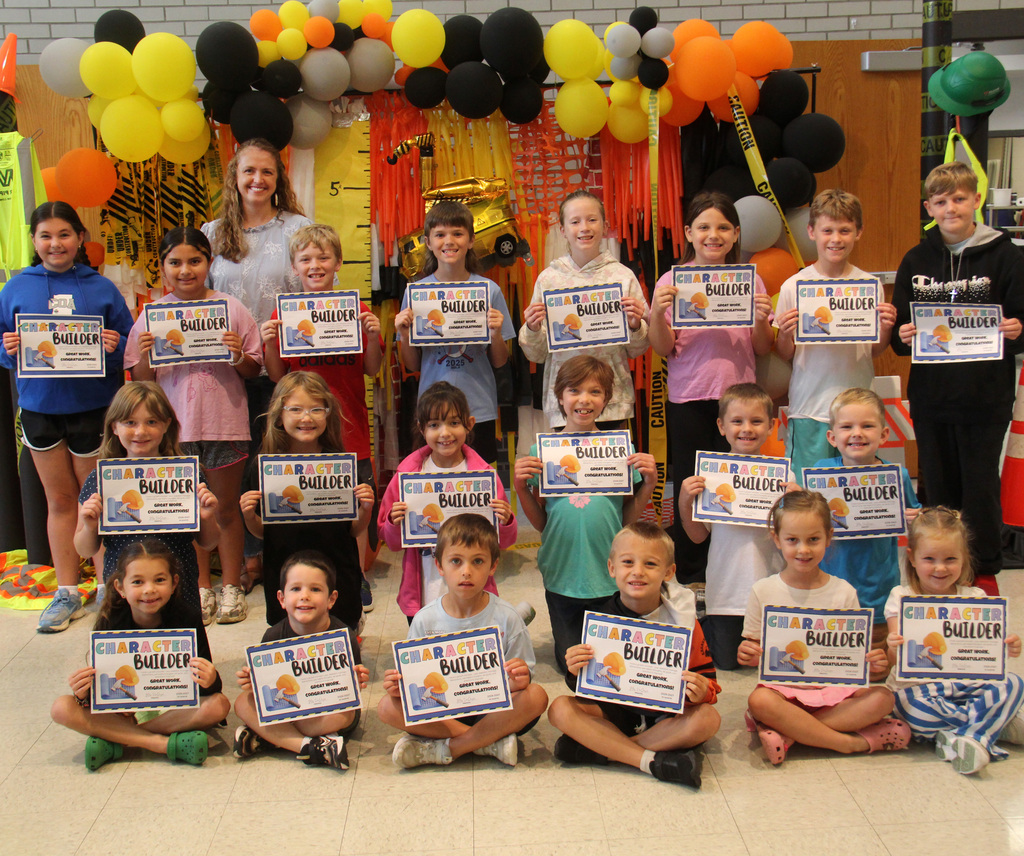 Students selected for the Character Builder of the Week, stand posing for a photo holding their certificates with the Principal! There are construction decorations behind them for a backdrop.