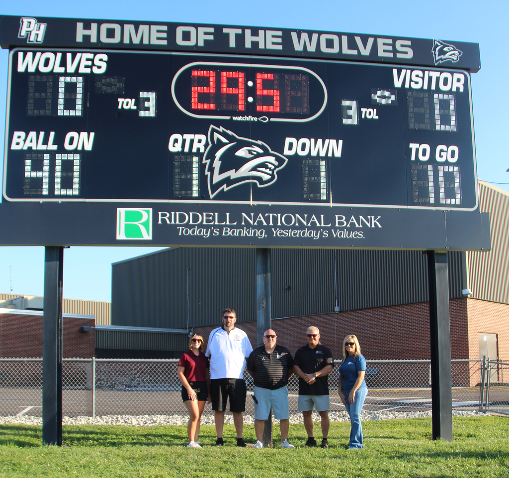3 individuals from Riddell National Bank and the principal and the athletic director at the ribbon cutting ceremony posing for a photo underneath the New Scoreboard.