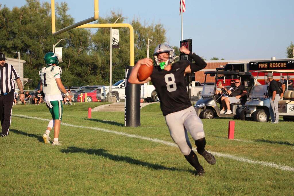 A Parke Heritage football player wearing number 8 runs toward the end zone carrying the football and raising his free arm. A defender in a white and green uniform follows behind him. The play takes place near the goal line, with the goalpost, officials, team vehicles, and spectators visible in the background.