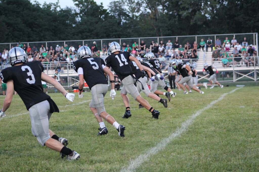 Parke Heritage football players in black jerseys, silver helmets, and gray pants move downfield during a play. Players wearing numbers 3, 5, and 14 are visible in the foreground, with additional teammates running toward the right side of the field. Spectators watch from the bleachers in the background.