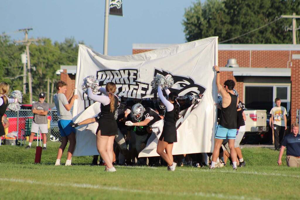 Parke Heritage football players run through a large white banner featuring the team name and wolf logo as cheerleaders hold pom-poms and players emerge onto the field.