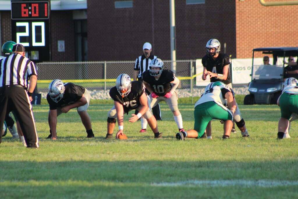 Parke Heritage football players in black jerseys and silver helmets line up against players in white and green uniforms before a play. A quarterback stands behind the center preparing to take the snap, while officials and players are positioned across the field. A scoreboard in the background shows 6:01 remaining and a score of 20.