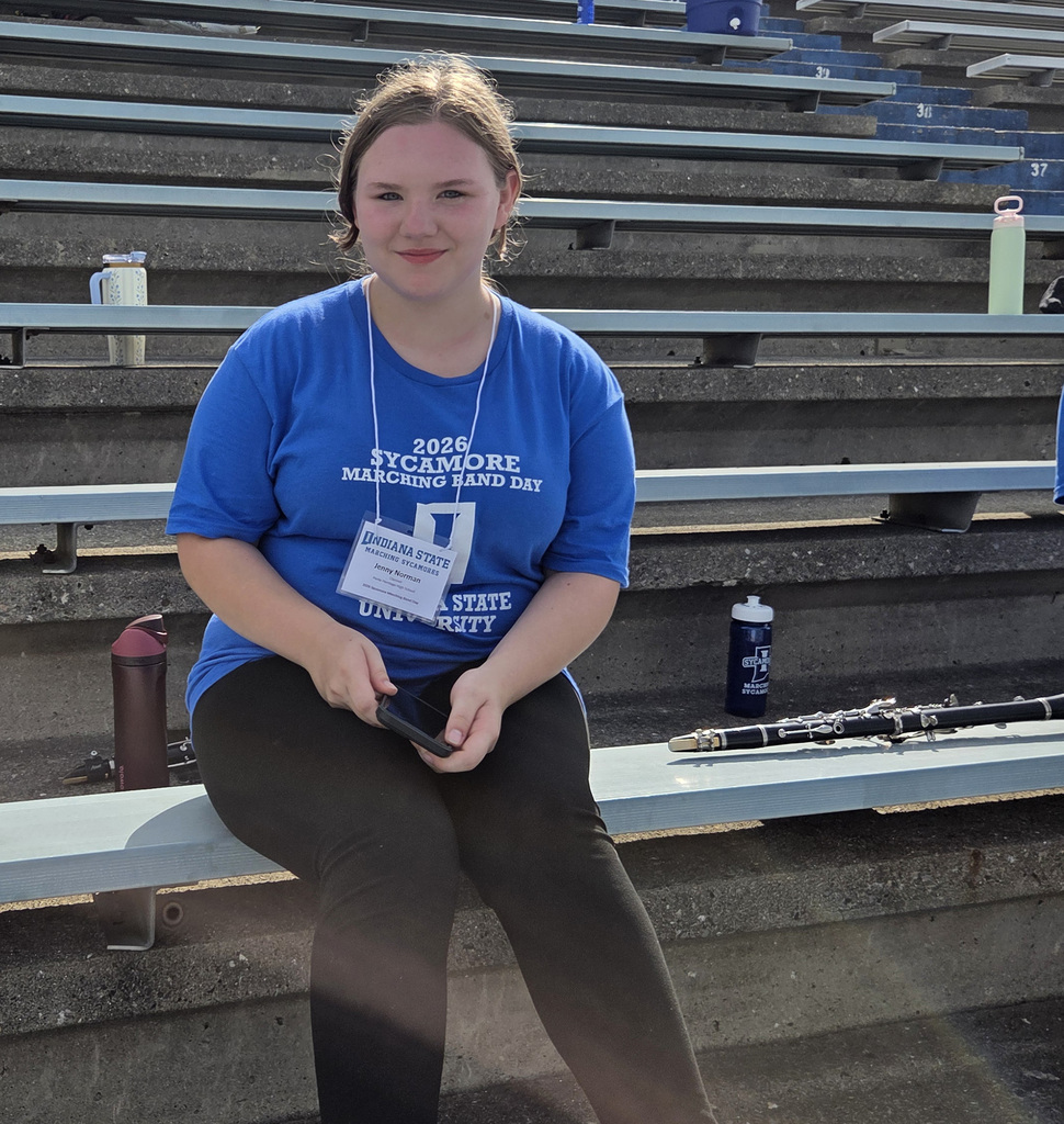 A student sits in the bleachers holding a phone and wearing a blue 2026 Sycamore Marching Band Day shirt and event badge. A clarinet rests on the bleacher nearby.