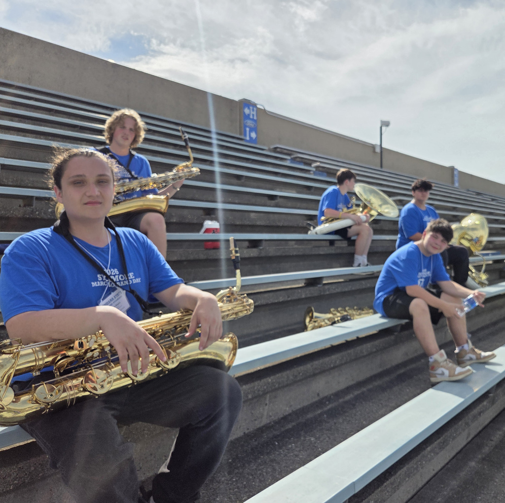Several students wearing blue Sycamore Marching Band Day shirts sit in the bleachers with their musical instruments, including saxophones and large brass instruments.