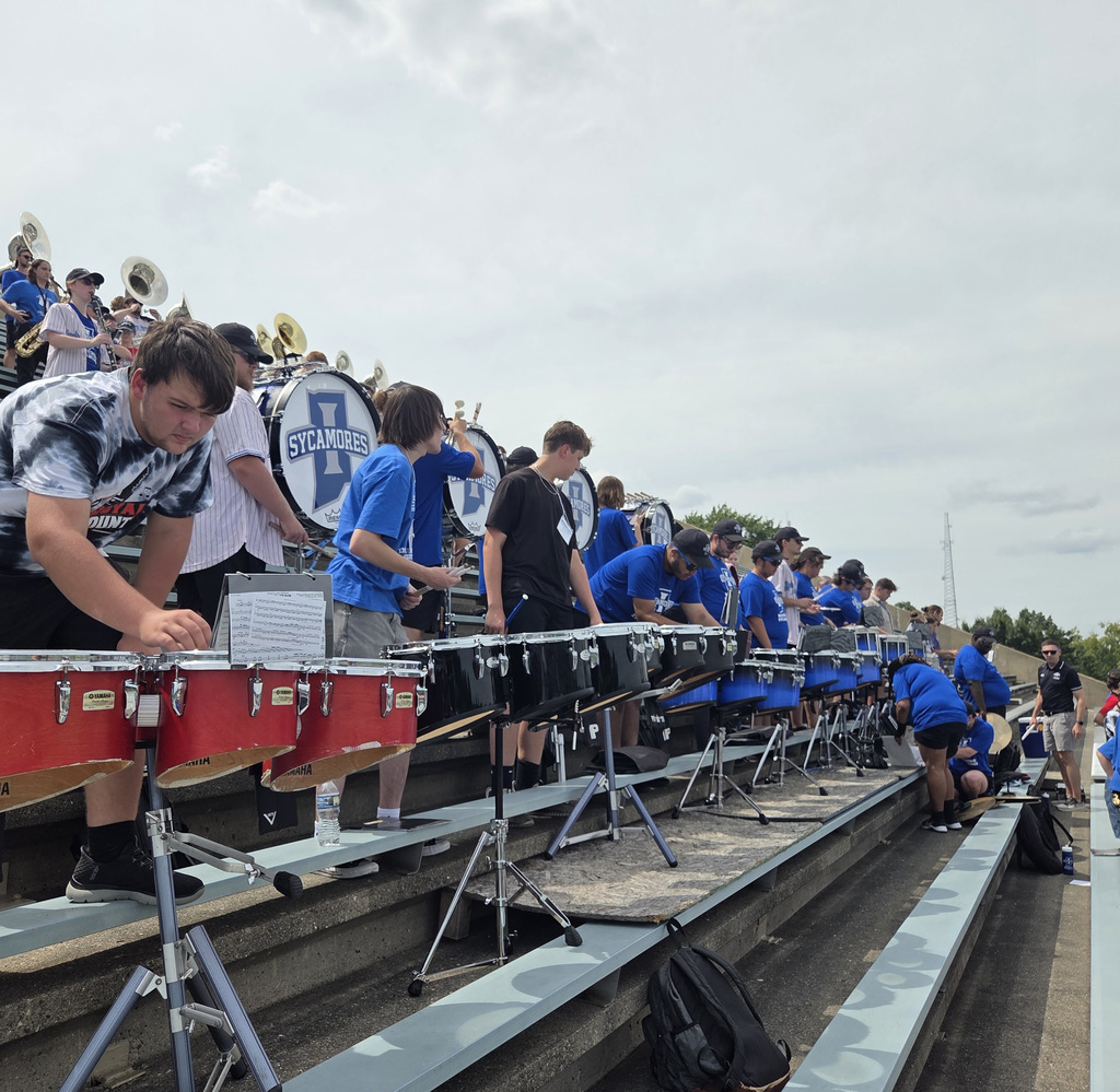 Marching band students rehearse with percussion instruments in the bleachers. A line of students plays drums while additional band members and large bass drums are visible behind them.