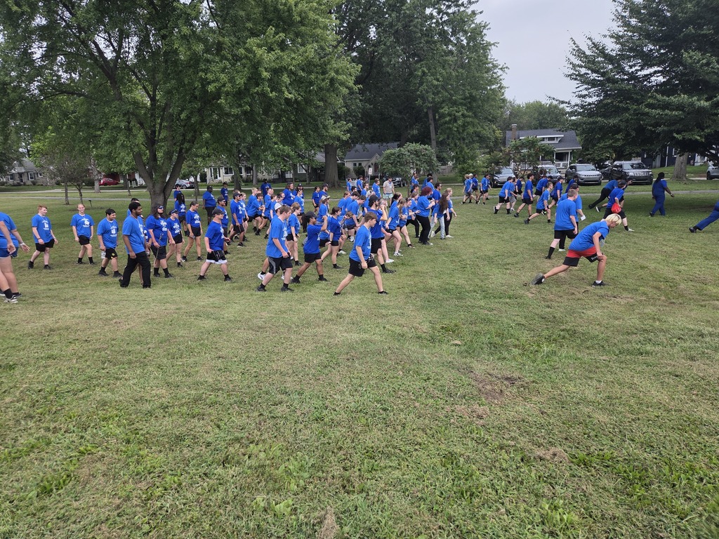 Marching band students participate in an outdoor group activity on a grassy field, moving together across the field while wearing blue Sycamore shirts.