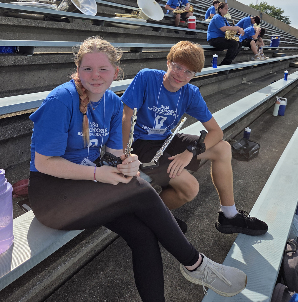 Two students sit together in the bleachers holding flutes and smiling for the photo. Other marching band students and instruments are visible in the background.