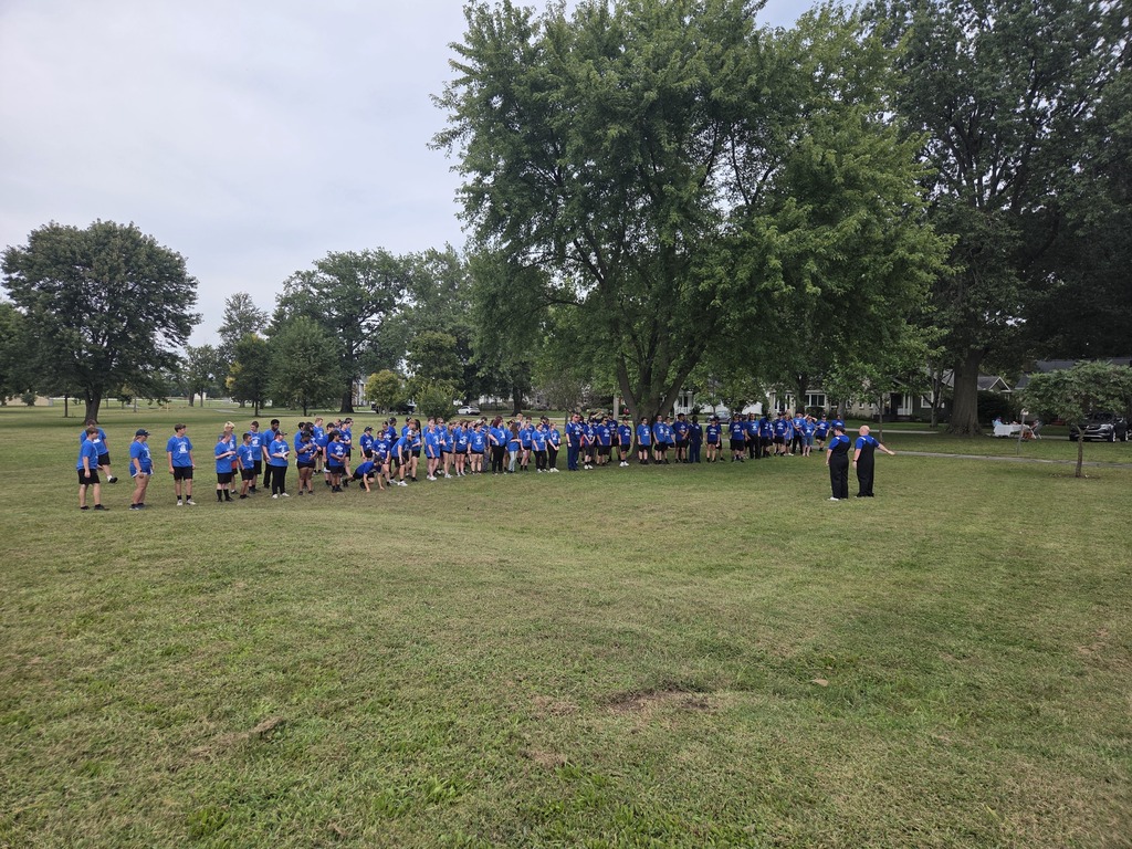 A large group of marching band students wearing blue shirts gathers on a grassy field during an outdoor band activity.