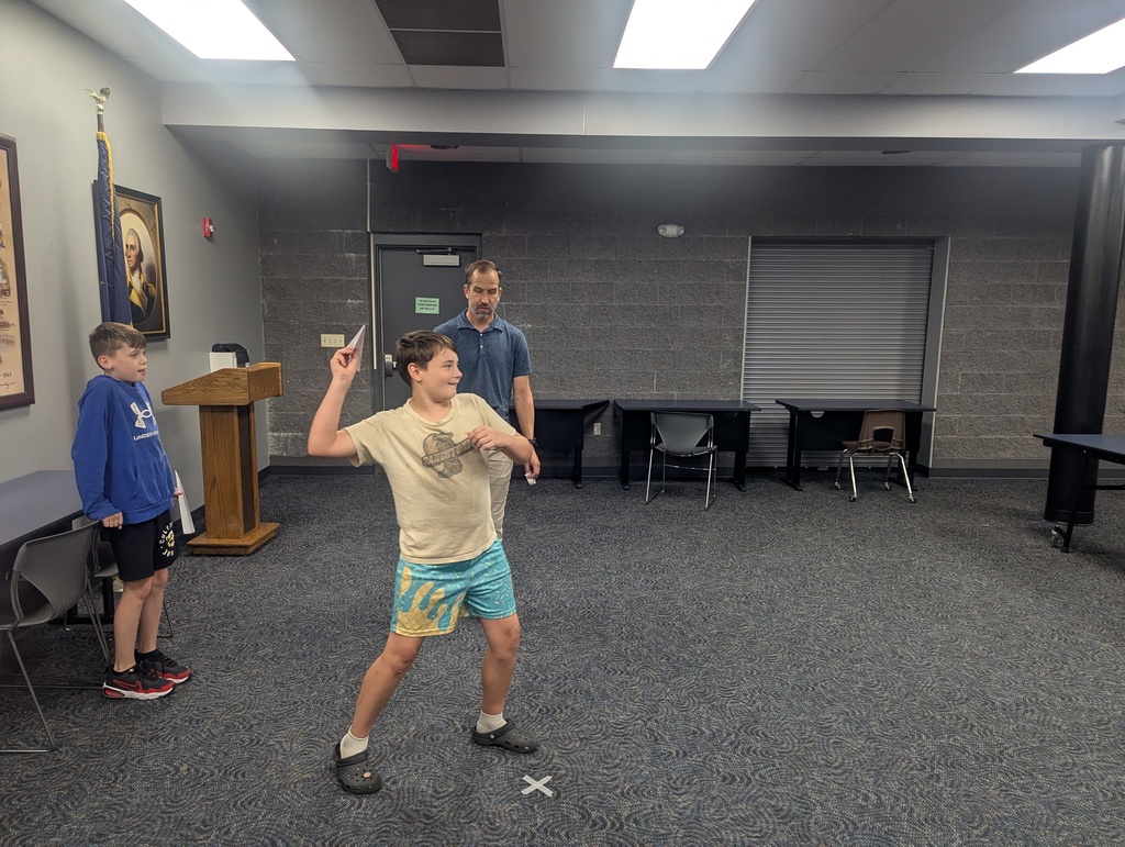 One student getting ready to fly his paper airplane while another student from his group and the teacher watch.