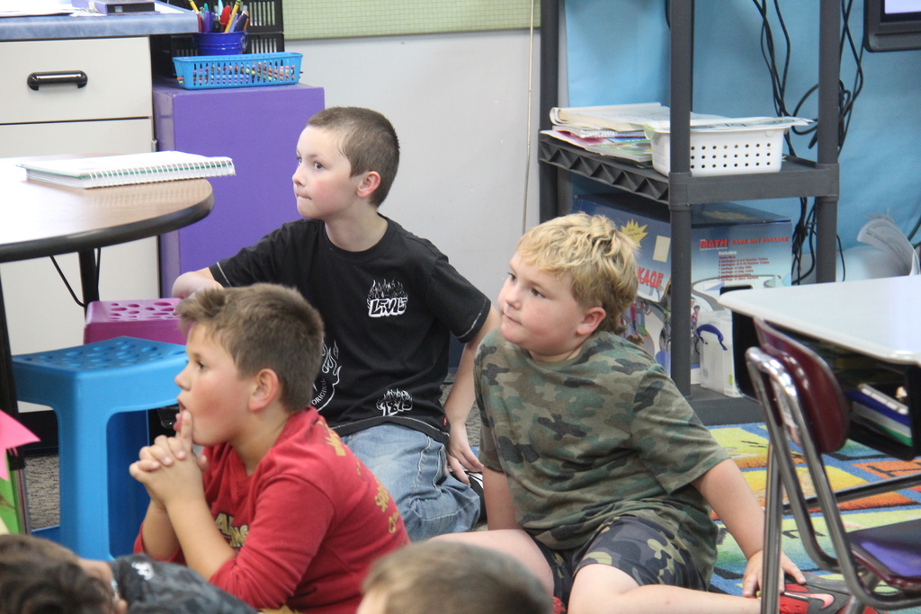 Students sitting on the floor while their teacher reads a book to them. 