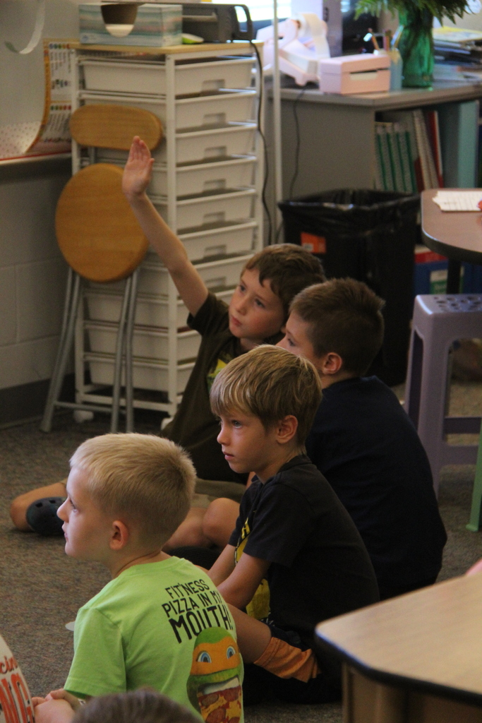 4 students sitting on the floor listening to their teacher. One student is raising his hand to ask a question. 