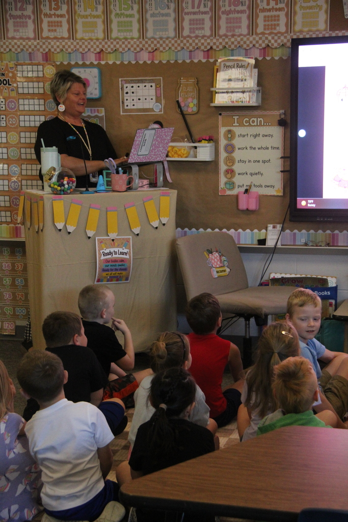 Students sitting on the floor listening while their teacher explains the screen in front of them. 