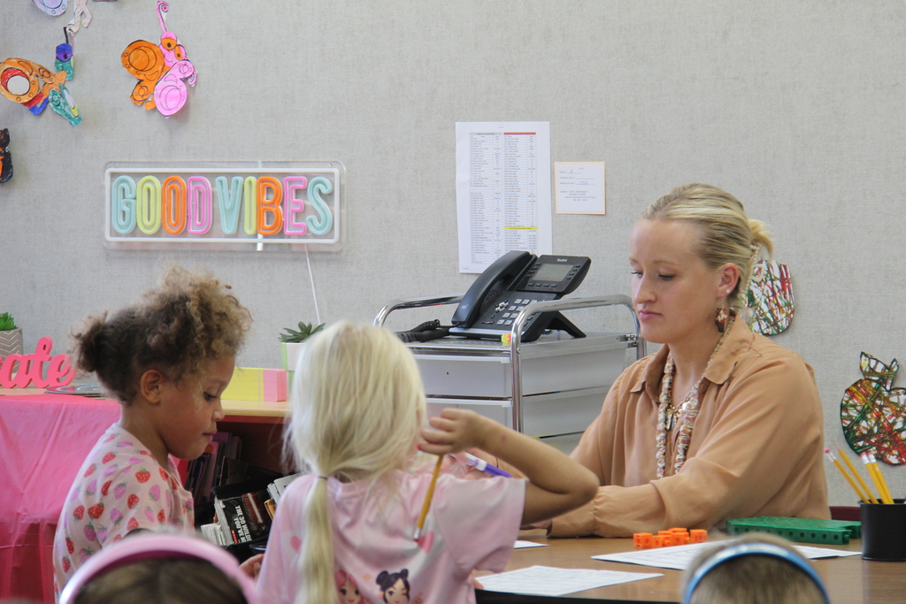 Teacher sitting at the table with a couple of students across from her while she helps them with their work. 