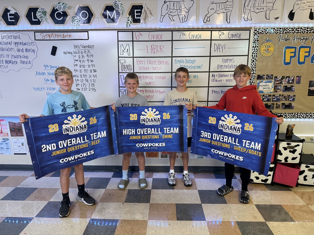 Four young livestock judging team members stand together in a classroom holding three large blue Indiana Youth Expo team award banners. The awards recognize 2nd Overall Team in Beef, High Overall Team in Swine, and 3rd Overall Team in Sheep/Goats. Livestock diagrams, a classroom schedule, and a Parke Heritage FFA display are visible in the background.
