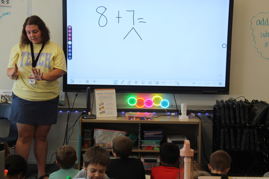 A second-grade teacher leads students through a math lesson involving addition on an interactive classroom display while students follow along.