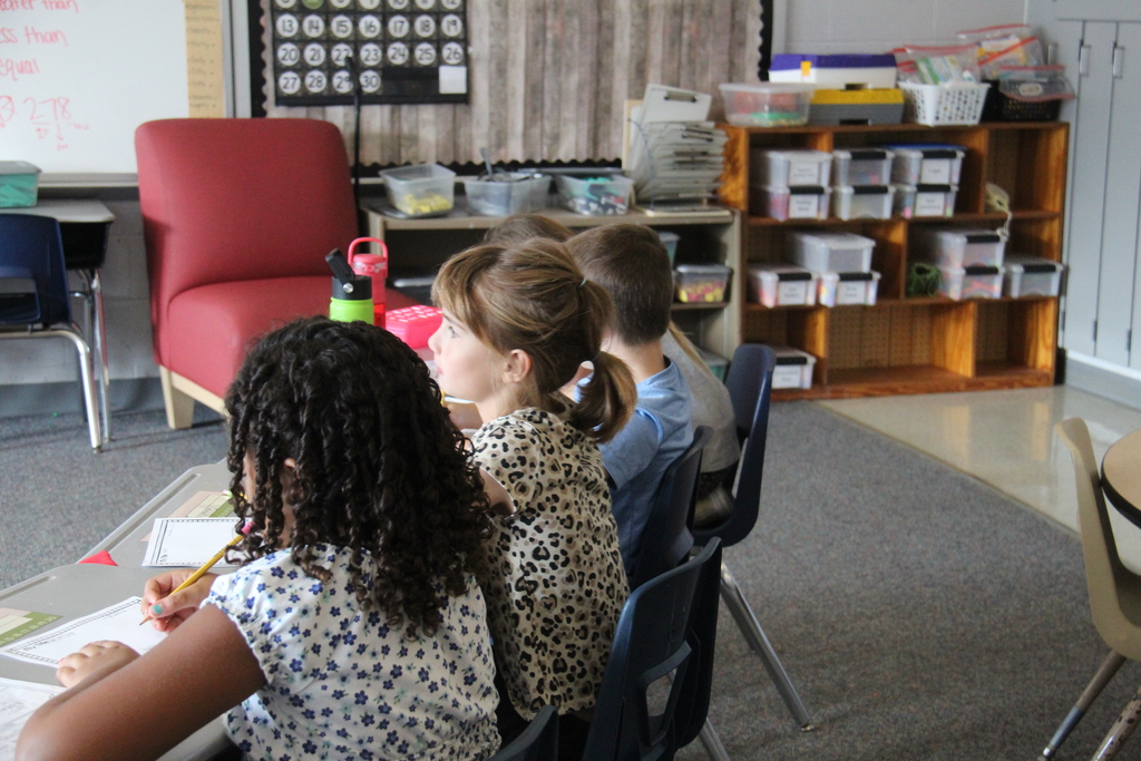 Three second-grade students sit together at a table, working on their classwork and listening to instruction.