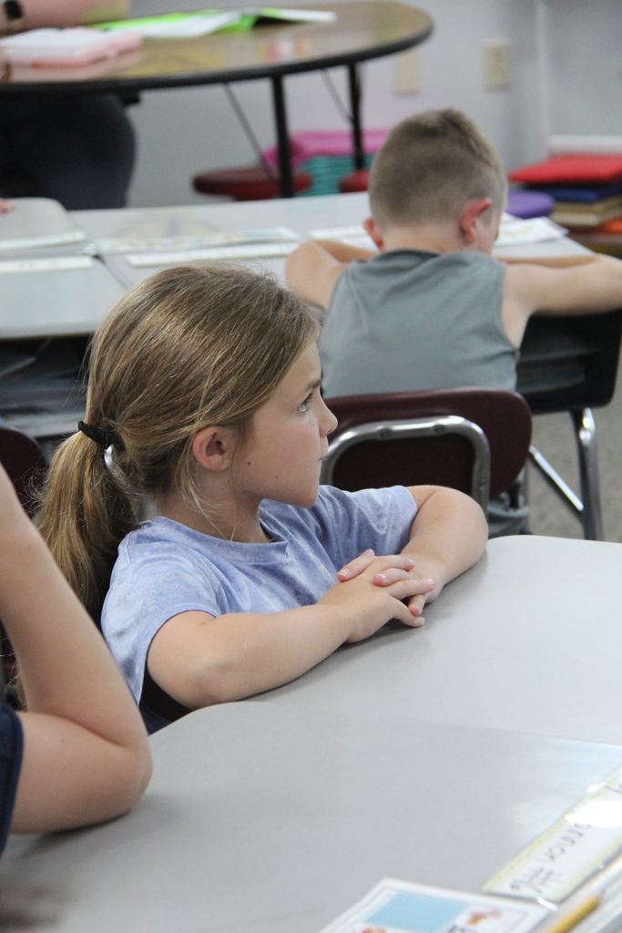 A second-grade student sits at a table with her hands folded while listening to the teacher; another student works nearby.