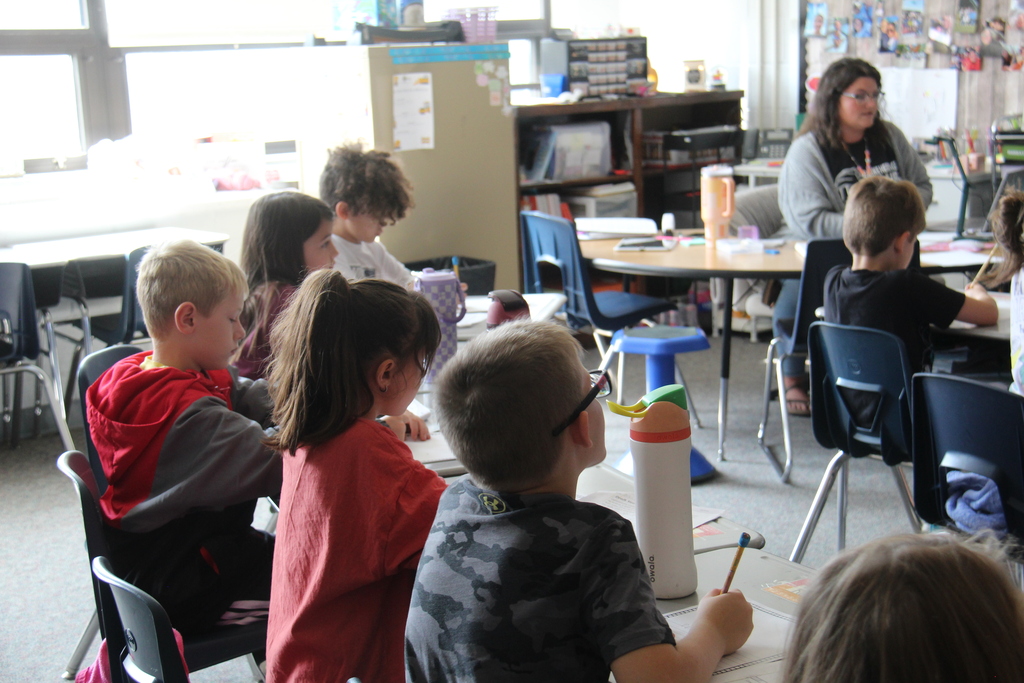 Second-grade students sit at classroom tables working on assignments while their teacher leads instruction from the front of the room.