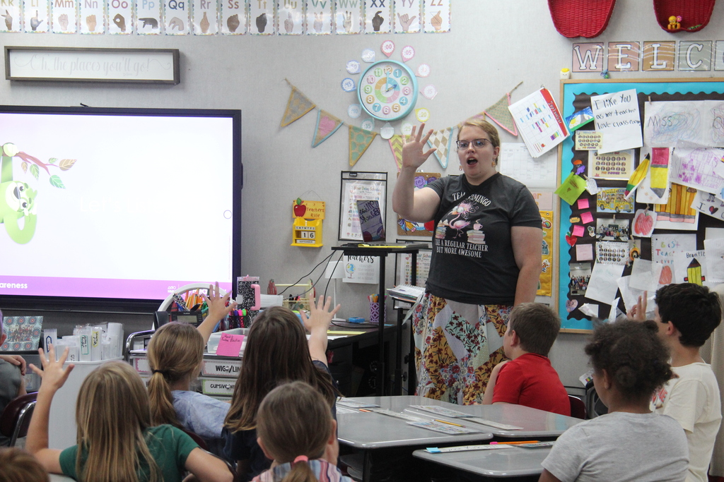 A teacher stands at the front of a second-grade classroom leading a lesson while students raise their hands and participate.
