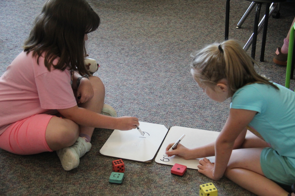Two second-grade students work together on whiteboards while sitting on the classroom floor, practicing writing or math skills.