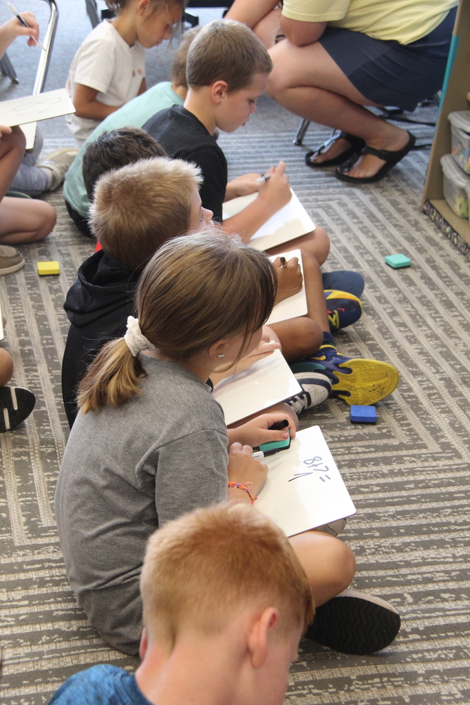 A teacher works with two second-grade students at a table as they complete a classroom activity using whiteboards.