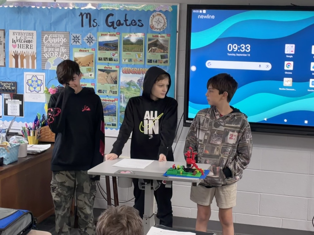 Three students stand at the front of a classroom presenting a colorful LEGO model. One student wears a black shirt and camouflage pants, another wears a black hoodie with a graphic on the front, and the third wears a gray patterned sweatshirt with shorts. A geography-themed bulletin board and interactive display are visible behind them.