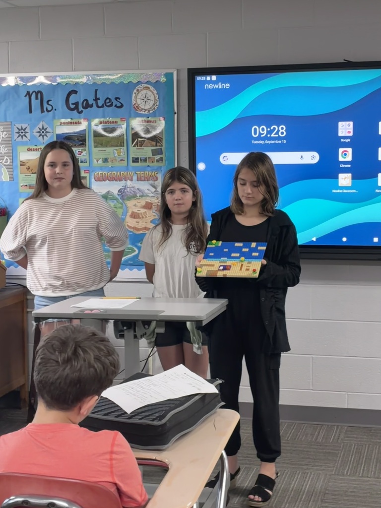 Three students stand at the front of a classroom with a LEGO model. One student holds the model, which features a blue surface and tan-colored sections. The students are standing near a classroom desk, with a geography-themed bulletin board and interactive display in the background.