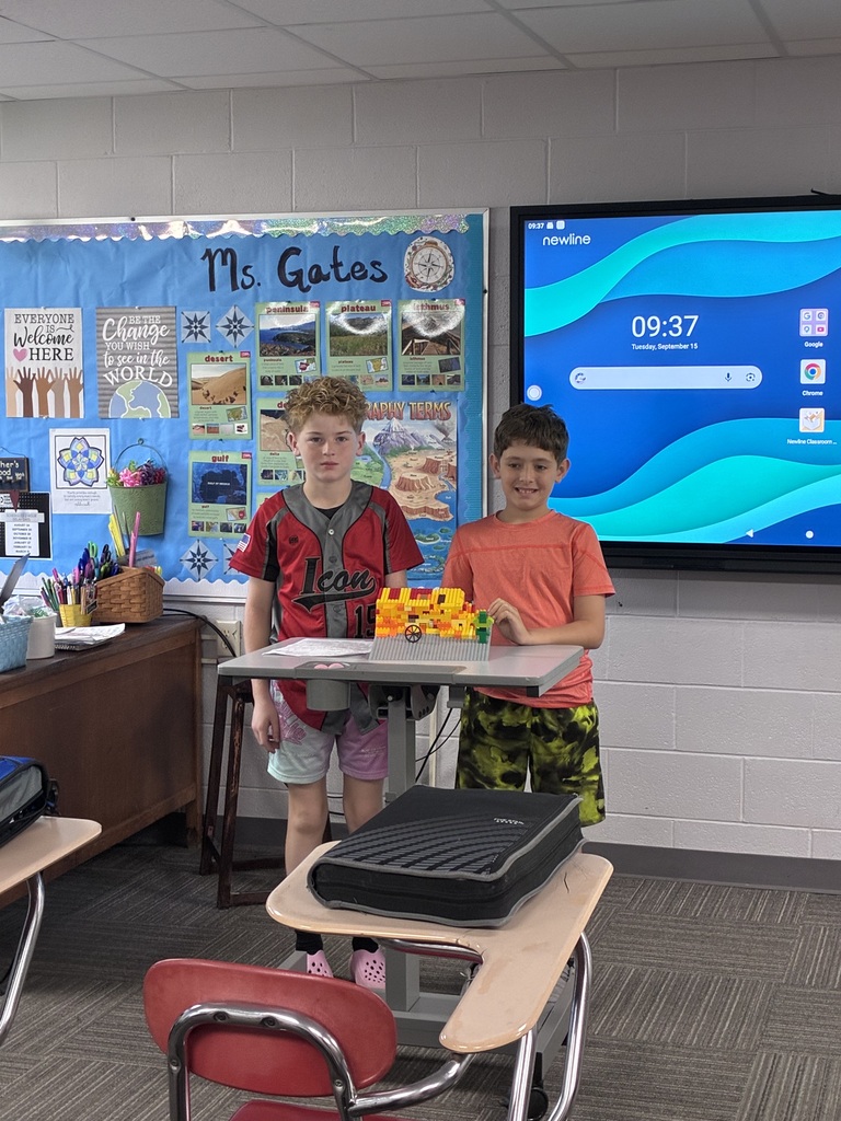 Two students stand at the front of a classroom beside a small LEGO model displayed on a desk. One student wears a red and black baseball-style shirt, another wears a coral shirt, and the third wears a gray patterned sweatshirt. A geography-themed bulletin board and interactive classroom display are visible in the background.