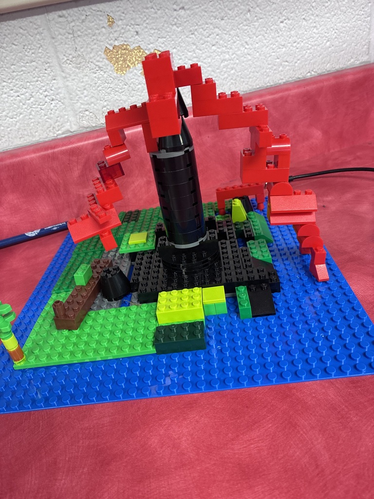 A close-up photo shows a colorful LEGO model built on a blue base. The model features a tall black-and-red structure surrounded by green areas, blue sections, and smaller LEGO details.