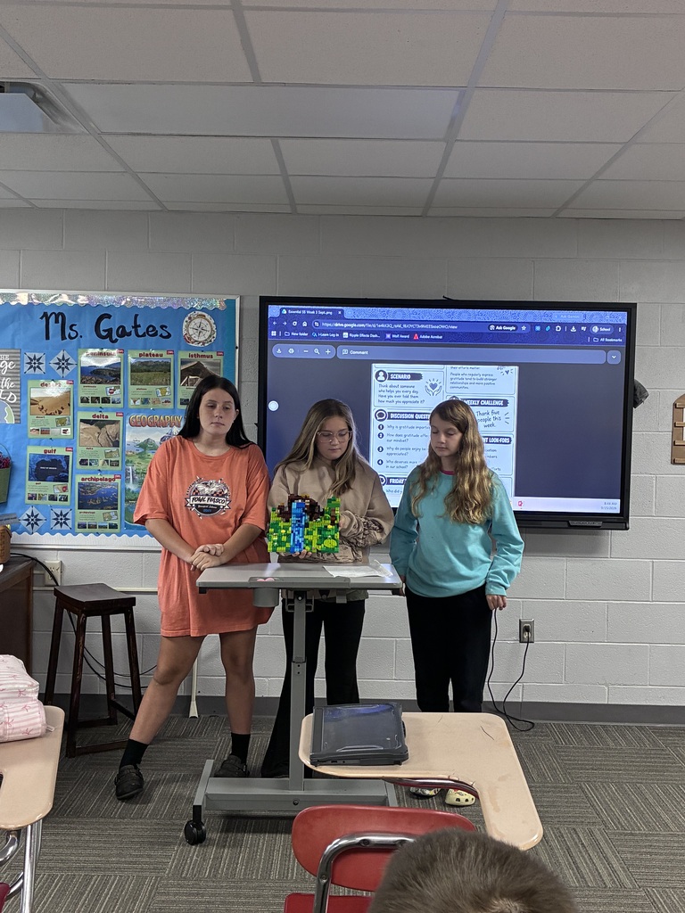 Three students stand at the front of a classroom, with two students holding a colorful LEGO model built on a rectangular base. The model includes blue, green, yellow, and brown LEGO pieces arranged into a detailed landscape. Behind the students is a geography-themed bulletin board labeled “Ms. Gates,” along with an interactive classroom display showing a digital worksheet.