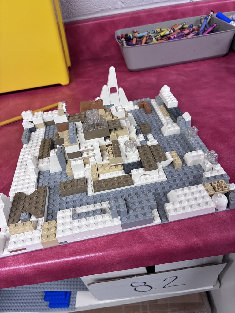 A close-up photo shows a LEGO model built primarily with white, gray, tan, and brown pieces. The model includes a detailed landscape with several raised structures and small transparent LEGO pieces. A handwritten number is displayed beneath the model.