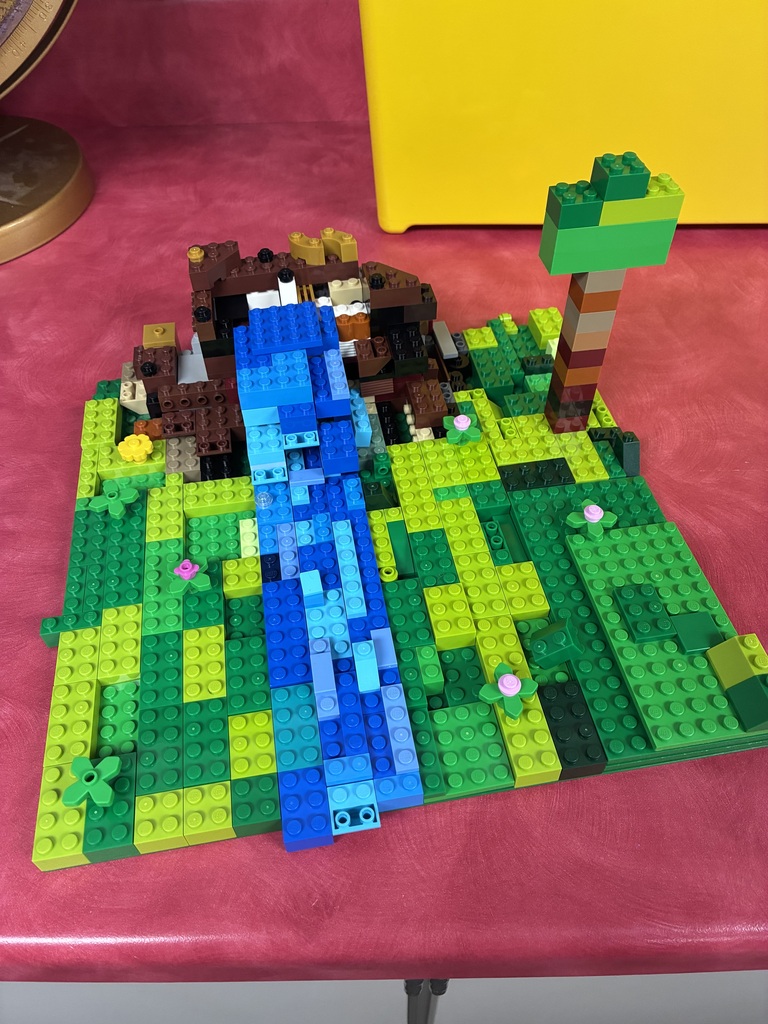 A colorful LEGO landscape model features a blue waterfall or river flowing through a green landscape. The scene includes a brown structure, a tall multicolored tree, flowers, plants, and other landscape details.