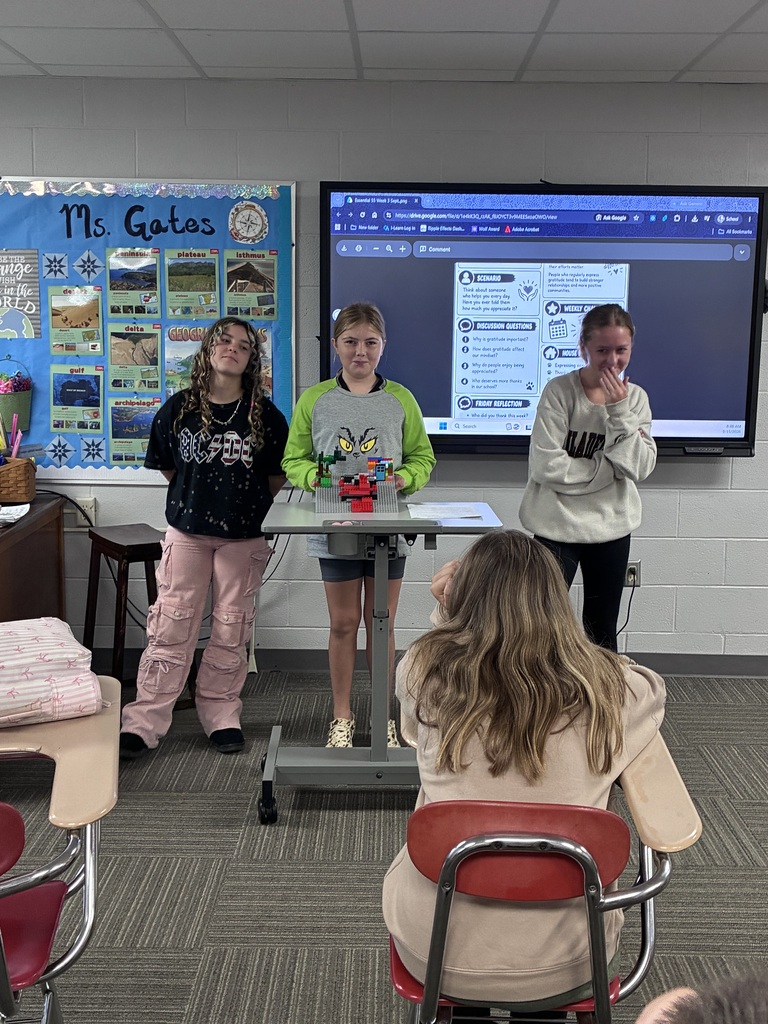 Three students stand together at the front of a classroom while presenting a colorful LEGO model featuring a central structure. One student wears an orange shirt and shorts, another wears a tan shirt and dark pants, and the third wears a teal shirt and black pants. An interactive classroom display and geography-themed bulletin board are visible behind them.