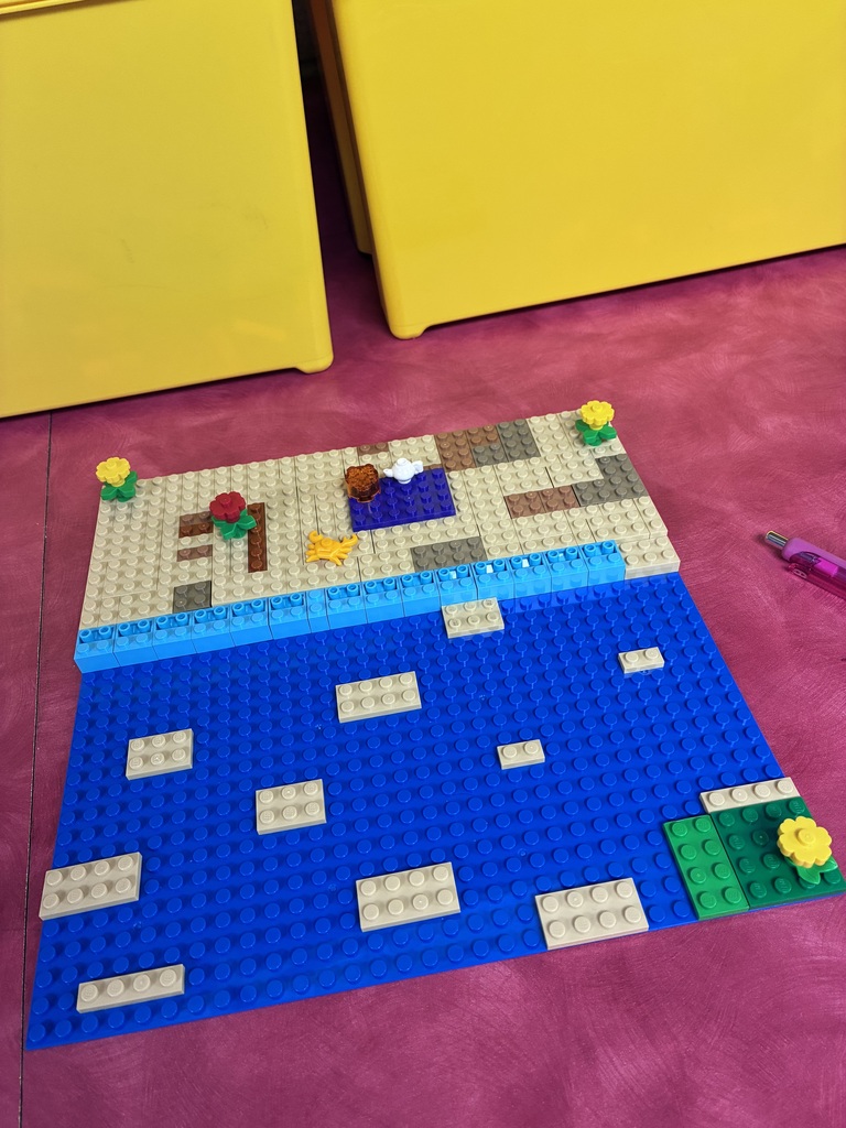 A LEGO landscape model features a large blue water area bordered by a sandy-colored beach. Small LEGO pieces represent flowers, plants, rocks, and other details along the shoreline.