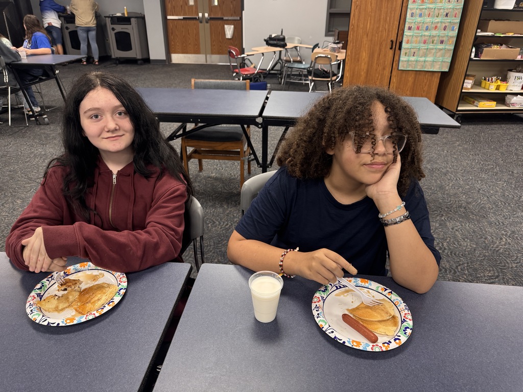 One student smiling for a photo with her fork in a pancake and another student is day dreaming with her fork also stuck in her pancake. 