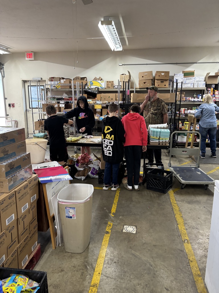 4 students packaging eggs at the food pantry with one adult supervising and another adult in the background working.
