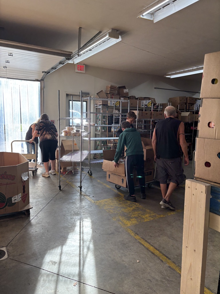Two adults in the background working, 2 students unpacking boxes and one adult supervising the students. 