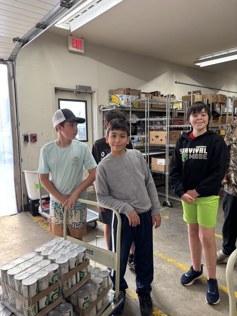 2 students leaning on a cart full of canned green beans while posing for a photo and one student in the background posing for the photo also.
