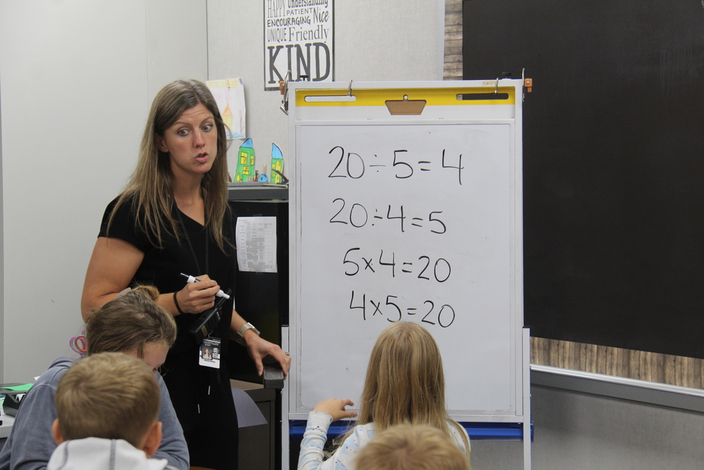 A teacher holding a dry erase marker after writing math problems on the white board and explaining the problems to the students sitting in front of her. 