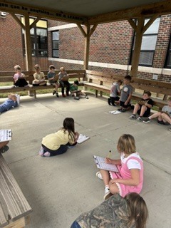 1st grade students all sitting around under the pavilion working on their math worksheets. 