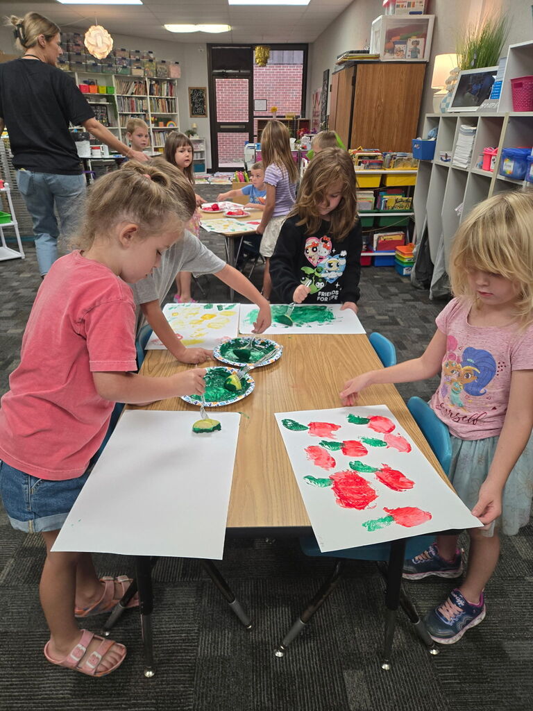 Four students standing around a table creating apple art using an apple and dipping it in paint and painting with it.  Other students at a table in the background doing the same thing. 