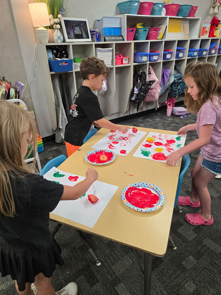 Three students standing around a table creating apple art using an apple and dipping it in paint and painting with it. 