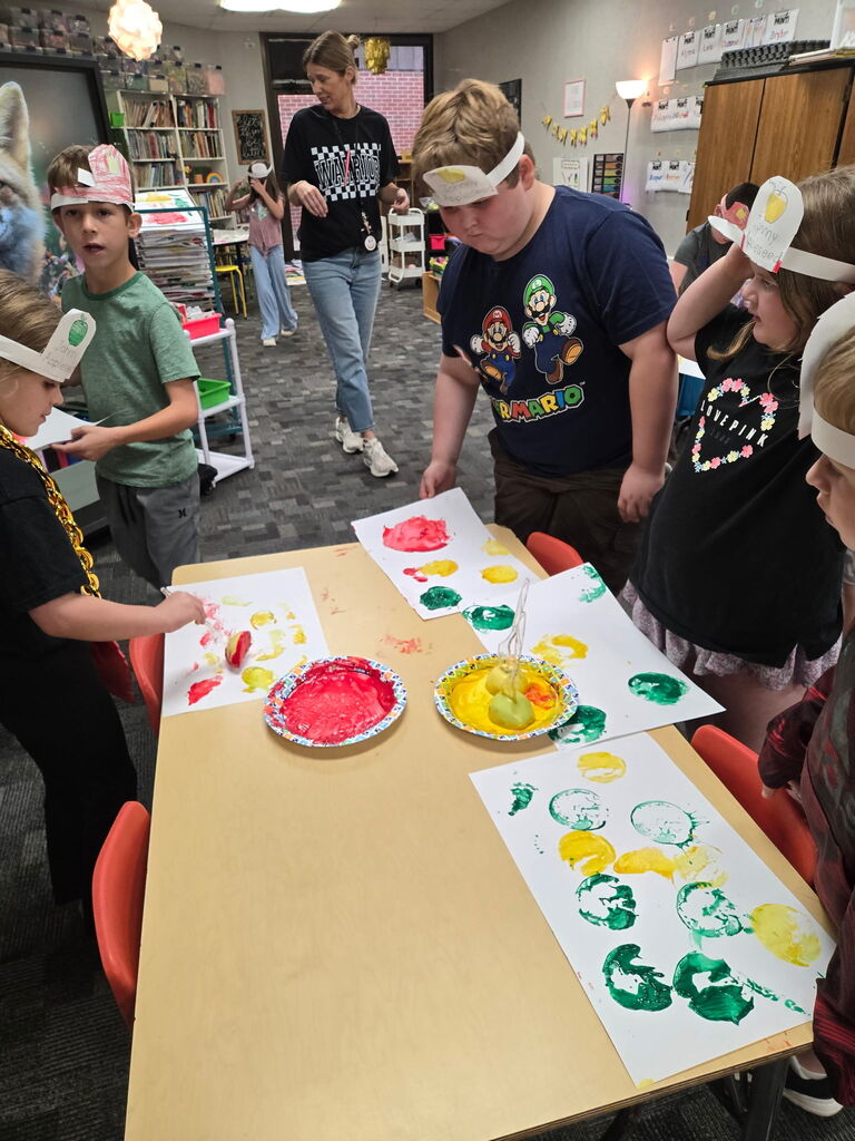 Four students standing around a table creating apple art using an apple and dipping it in paint and painting with it. 
