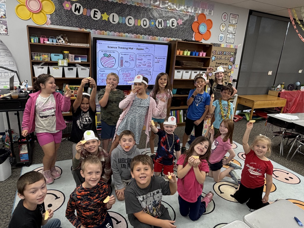 Several students sitting on the floor and several students standing in the back, facing the camera, showing off their apple smiles. 