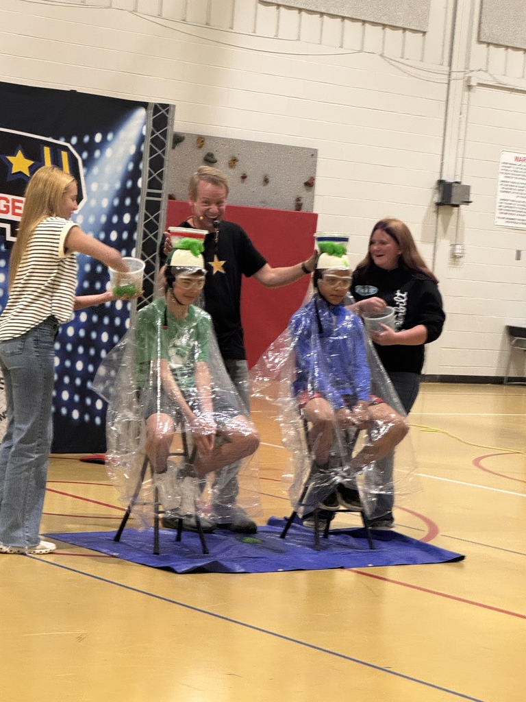 2 students sitting on stools with hand made hats that have a bowl on top with slime from the competition in the bowl. 