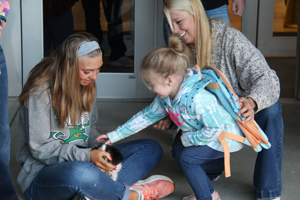 One of the students leans over to pet the puppy, held by an athlete while another teacher leans down with the student. 