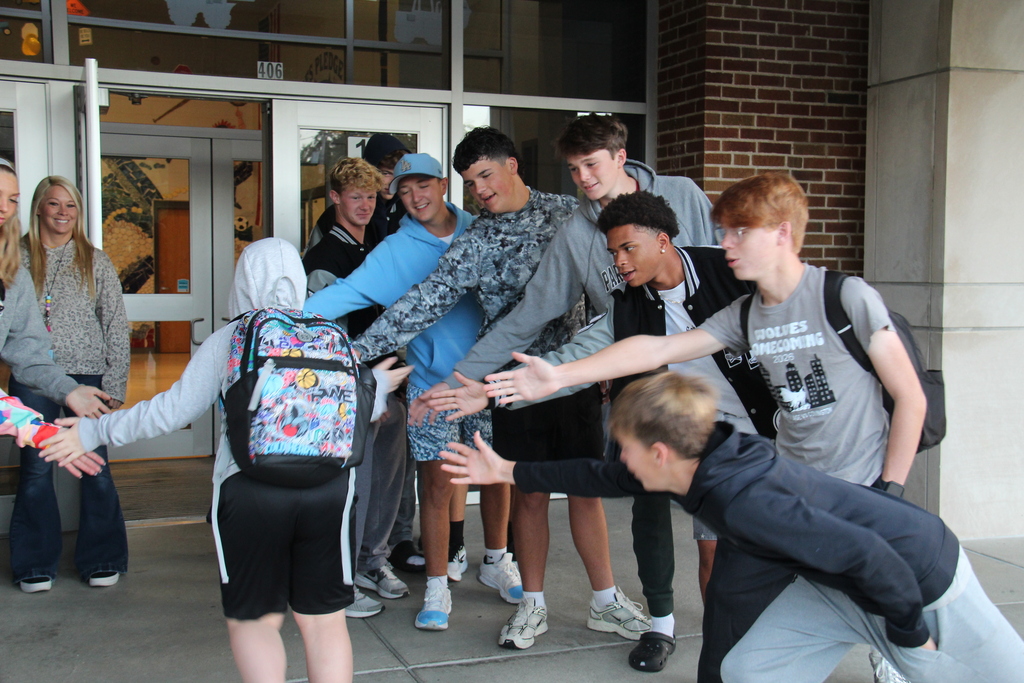 Members of the Tennis, Cross Country, and Golf teams line up along the sidewalk to five high fives to one of the students as he walks into school. 